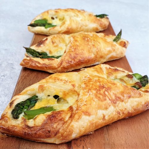 3 puff pastry asparagus wraps shown on a thin wooden board at a shallow angle. 